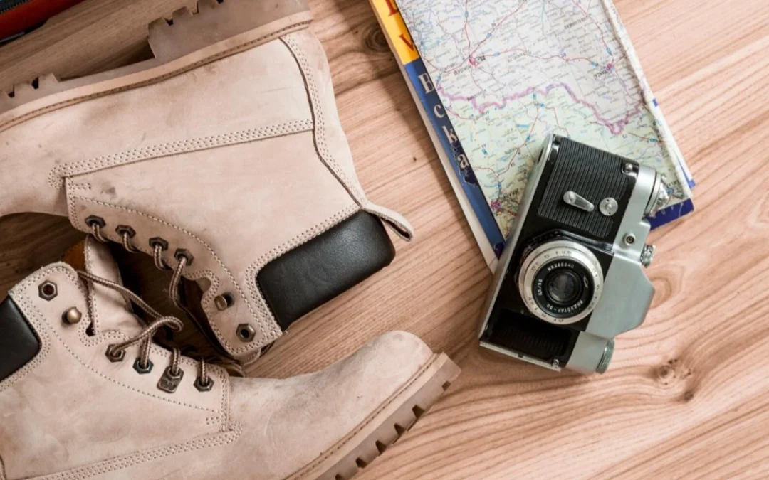Botas de senderismo color beige, cámara analógica vintage y mapa de viaje sobre una superficie de madera clara. Objetos que inspiran un viaje slow travel.
